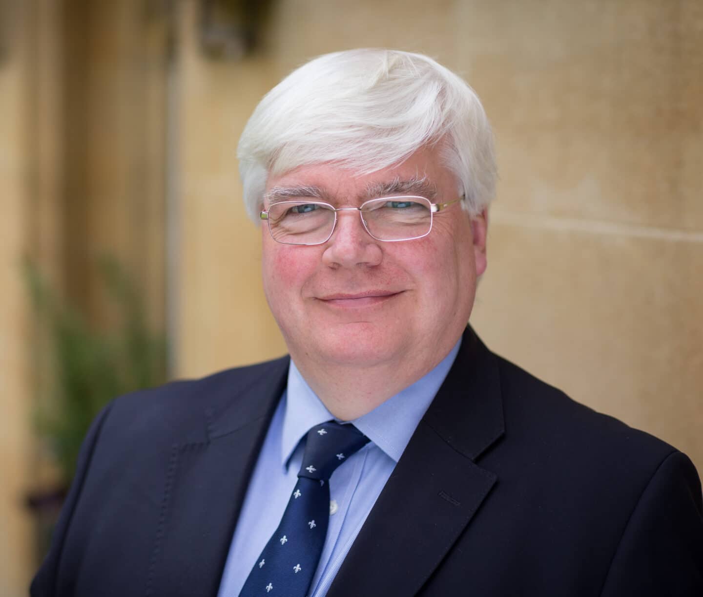 Portrait of David Vaux, with short white hair wearing glasses, a dark suit jacket, light blue shirt, and navy patterned tie, smiling slightly in front of a stone wall background.
