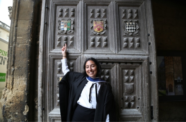Maria Murad in sub fusc standing in front of the carved Lincoln College front door, smiling and pointing to a carved crest above her.