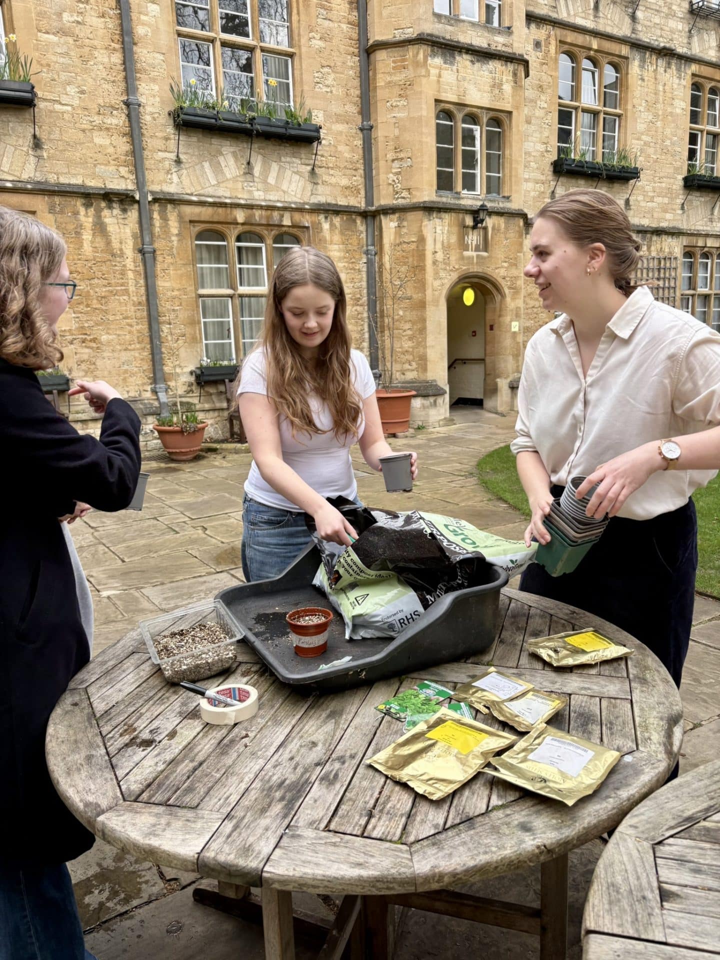 Staff and students take part in a seed-sowing activity in Lincoln College’s Grove Quad, filling plant pots with compost and preparing packets of seeds on a wooden outdoor table.