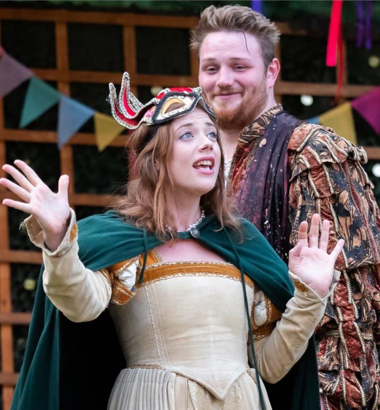 Beckis Cooper performing as Beatrice in Much Ado About Nothing during an outdoor theatre production. She wears an elegant cream and gold Renaissance-style dress with a green cloak and a decorative mask on her head. Her expressive gesture and animated facial expression convey confidence and wit. Behind her, a male actor dressed in rich period costume smiles, with colorful bunting strung in the background, suggesting a festive setting.