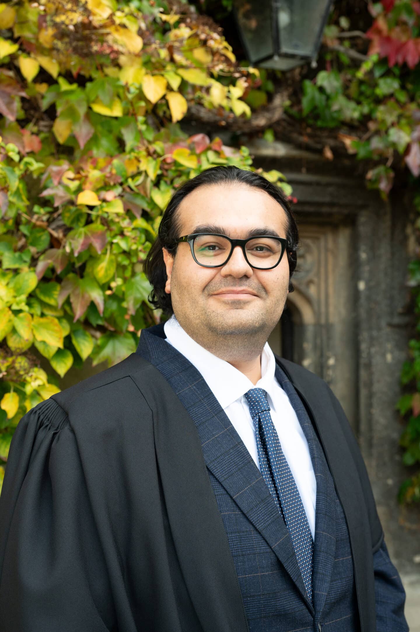 Mahan Ghafari standing in Front Quad at Lincoln College, Oxford, wearing a dark suit and tie, in front of a wall covered with green and yellow Boston ivy.